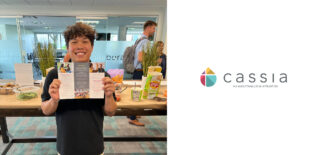Devon, the Diversity and Inclusion Intern at Cassia, smiles while holding a brochure during his farewell gathering at the Cassia Home Office, surrounded by snacks and drinks. The staff celebrated Devon's impactful contributions to promoting inclusivity within the community as he prepares to head off to school.