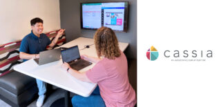 Devon and Jeri are seated at a table, each working on laptops. Devon, on the left, is smiling and gesturing towards a large screen on the wall displaying a spreadsheet and a website. Jeri, on the right, is looking at her laptop screen.