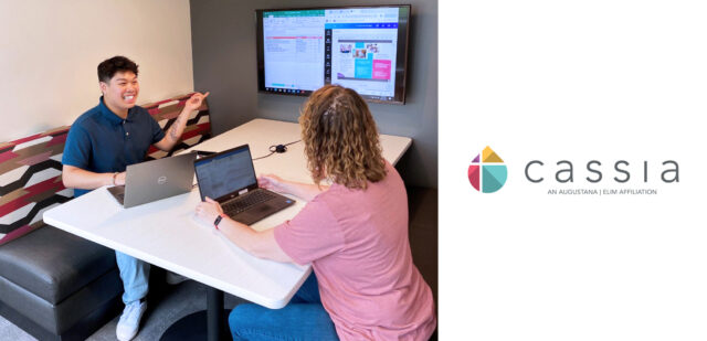 Devon and Jeri are seated at a table, each working on laptops. Devon, on the left, is smiling and gesturing towards a large screen on the wall displaying a spreadsheet and a website. Jeri, on the right, is looking at her laptop screen.