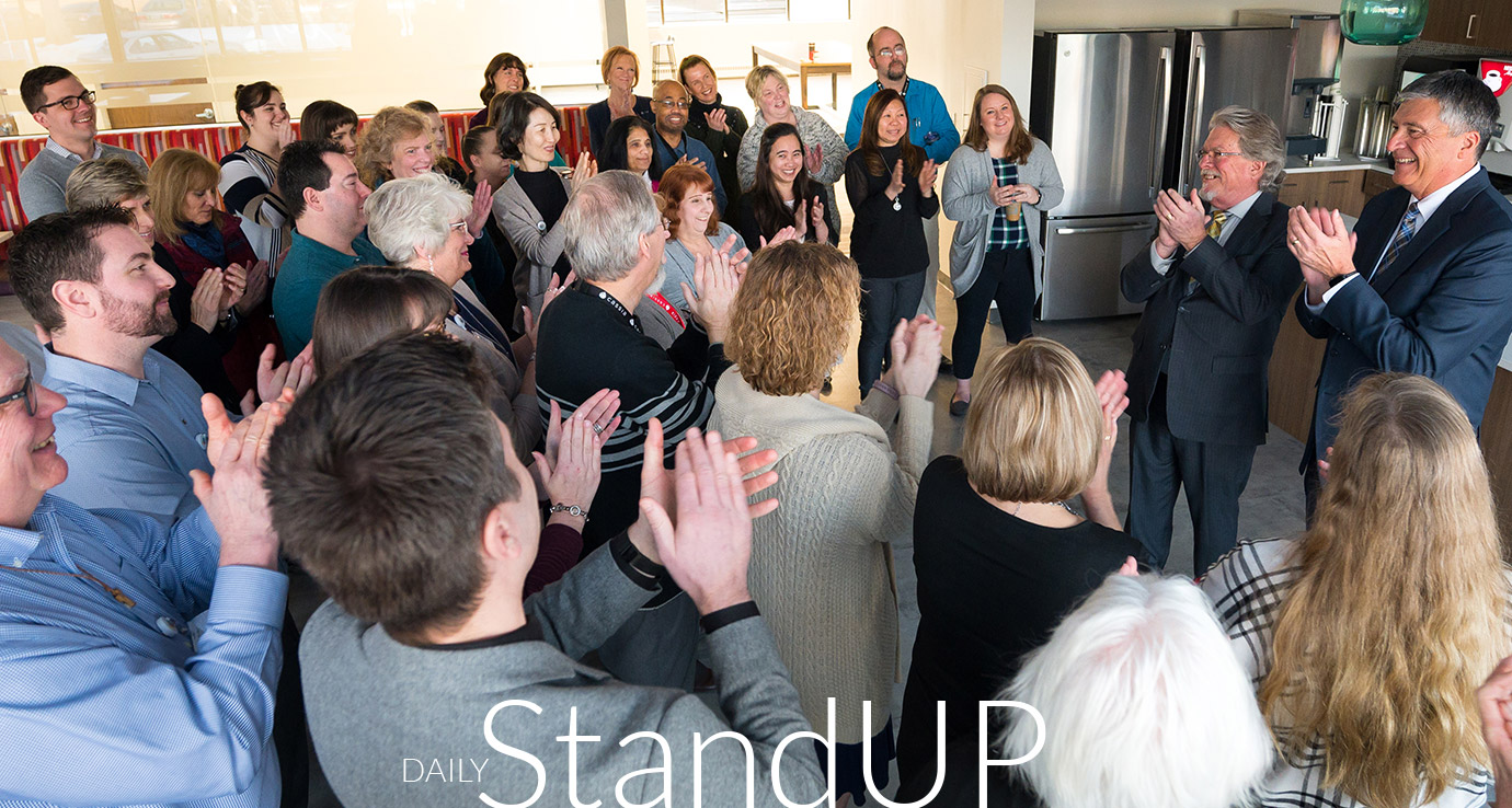 A group of Cassia employees gathered in a kitchen space, clapping and smiling during a Daily StandUP meeting. Two men in suits stand at the front, leading the discussion.