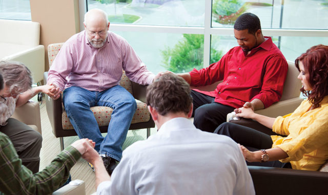 A diverse group of people sitting in a circle, holding hands in prayer inside a bright, welcoming space.