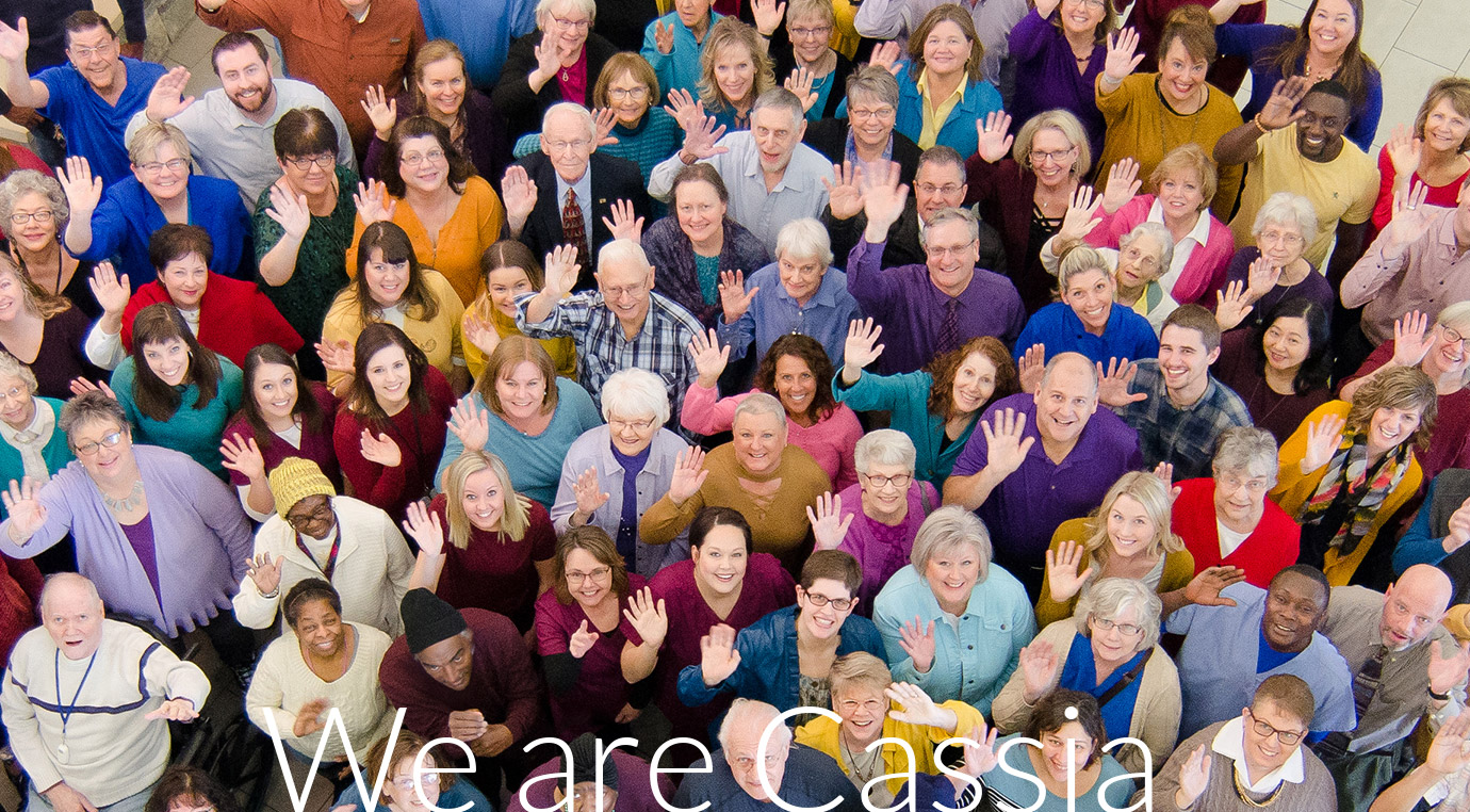 A large, diverse group of Cassia team members and community members smiling and waving at the camera, dressed in colorful attire. The words "We are Cassia" are overlaid on the image.