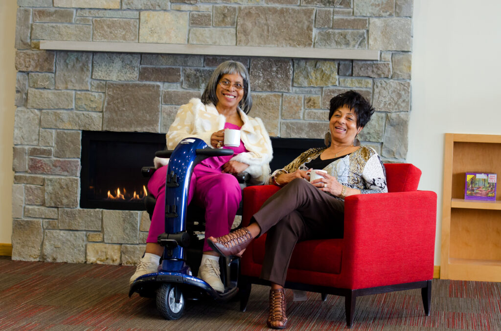 Two senior women sitting by a fireplace in an assisted living community, smiling and enjoying coffee together.