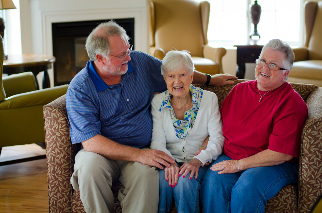 A senior woman sitting with two family members, laughing and sharing a moment in a cozy living room in an assisted living community. 