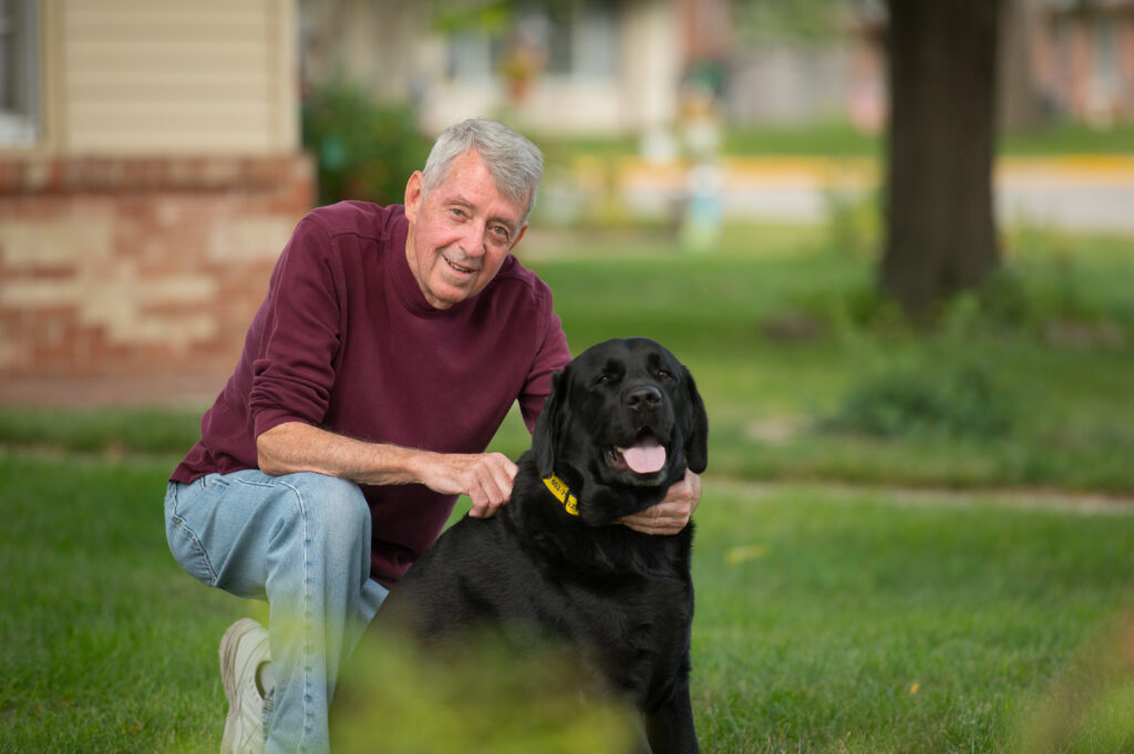 Older man sitting on the grass, petting a large black dog outside of independent living community. 