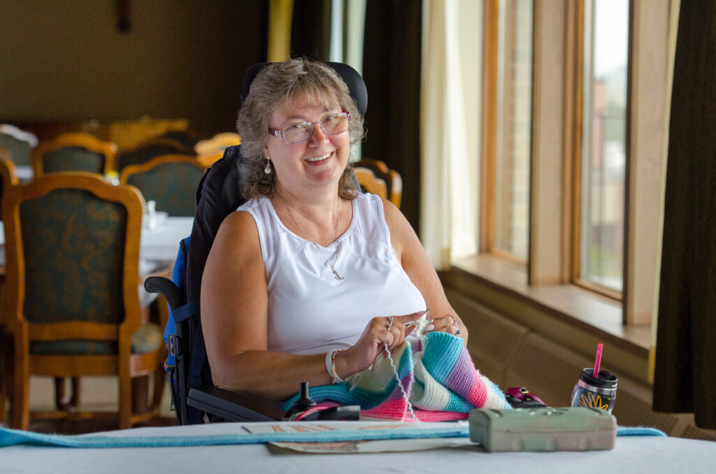 A senior woman in an assisted living community knitting at a table, smiling with natural light coming through large windows.