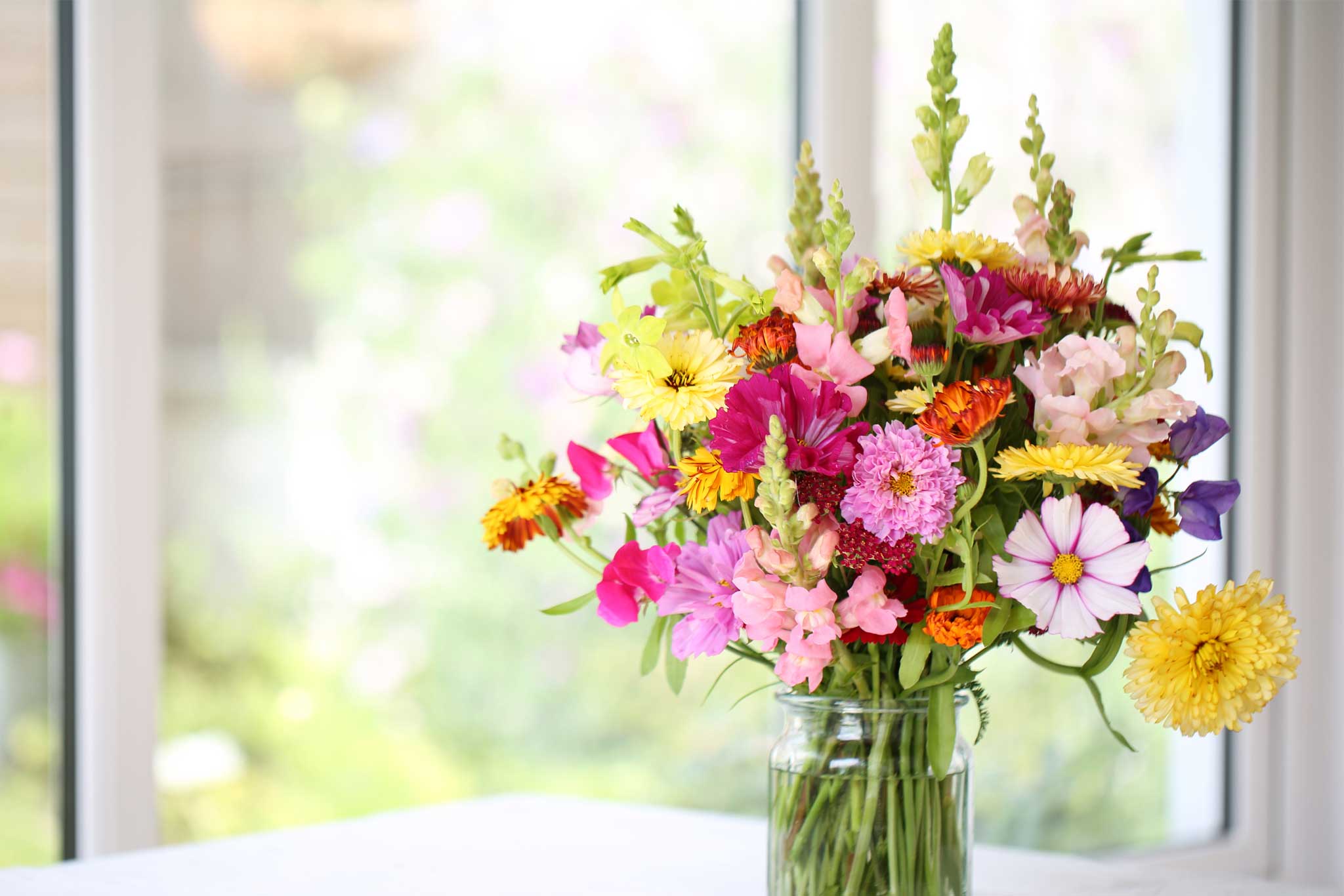 Colorful bouquet of mixed flowers in a glass jar, displayed on a white table with a bright window