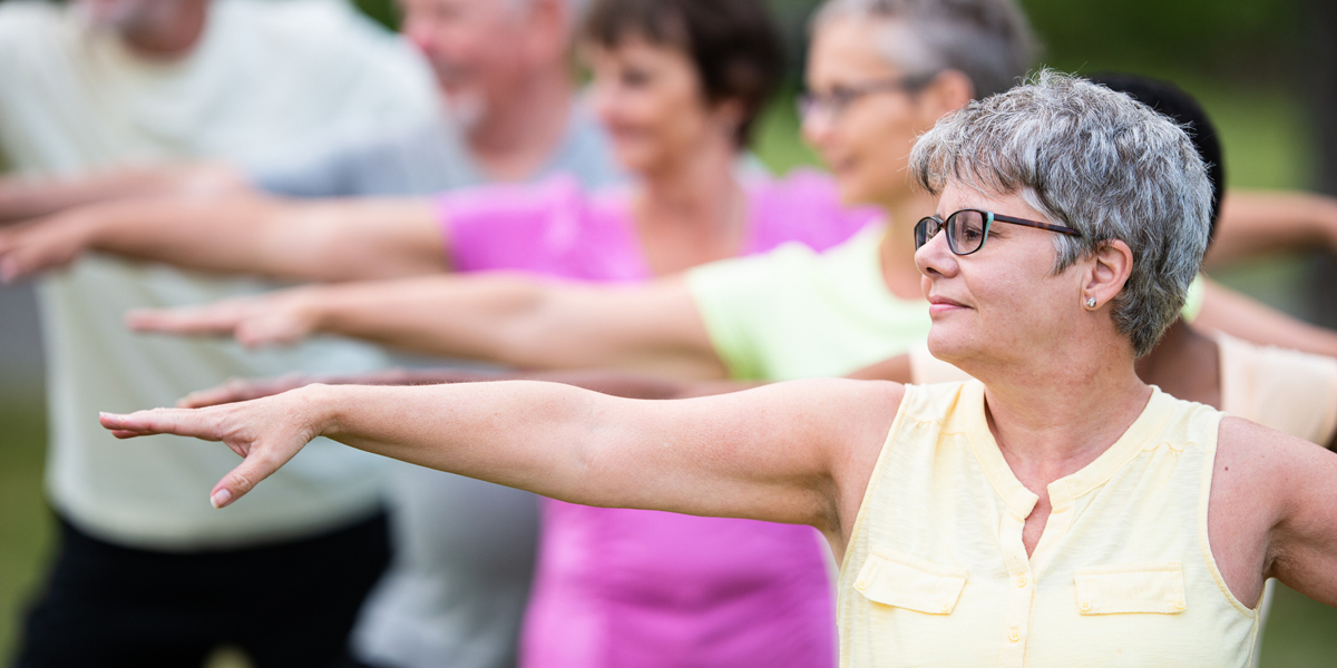 A group of multi-ethnic seniors gather together outside in a park to stretch during a fitness class. Each one is standing out on the grass with both arms stretched out horizontally. They are each dressed in comfortable active wear and smiling as they enjoy the class and the fresh air.