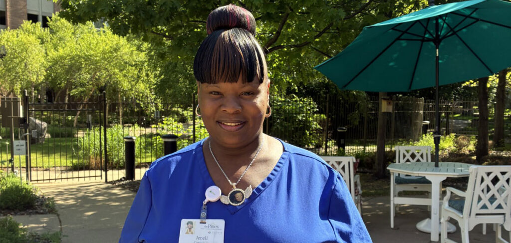 Caregiver at The Pines senior living community standing outdoors in a landscaped courtyard with patio seating and greenery, wearing blue scrubs and a name badge.