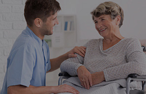 Caregiver sitting beside an adult using a wheelchair in a care setting, representing support programs that help bridge the financial gap for residents in need at Cassia