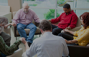 Group of adults seated in a circle holding hands in a shared gathering space, representing spiritual care and chaplain support at Cassia