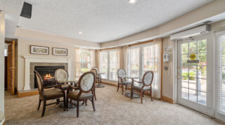 Comfortable seating area with small tables, chairs, fireplace, and large windows near an exterior door at The Pines of Richfield in Richfield, Minnesota