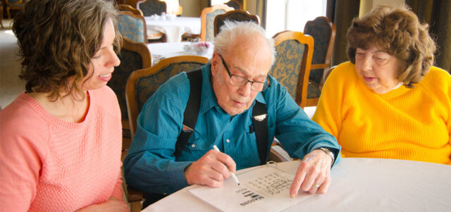 Three adults seated at a table working on a word or puzzle activity together in a dining area, representing care and engagement like The Pines of Richfield in Richfield, Minnesota