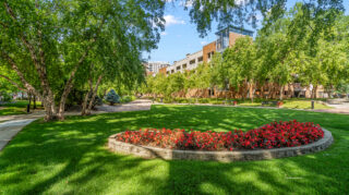 Landscaped outdoor garden with green lawn, curved flower bed, and mature trees surrounding The Pines of Richfield building in Richfield, Minnesota