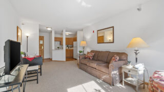 Apartment living room with sofa, side tables, lamps, dining table, and kitchenette in an open floor plan at The Pines of Richfield in Richfield, Minnesota