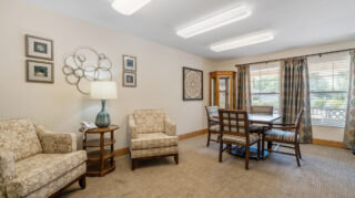 Lounge area with upholstered chairs, side table, and a table with seating near large windows at The Pines of Richfield in Richfield, Minnesota