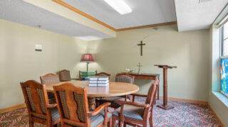 Small prayer room with round table, chairs, cross on the wall, and stained glass-style window at The Pines of Richfield in Richfield, Minnesota