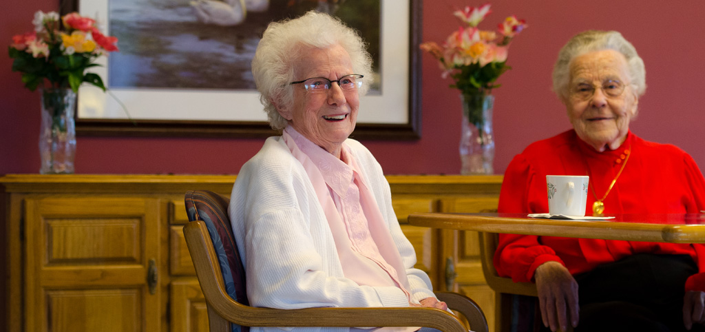 Two older adults seated at a table with a coffee cup in a cozy common area, representing social living like The Pines of Richfield in Richfield, Minnesota