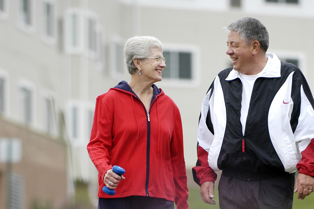 Two older adults walking outdoors together near a residential building, representing an active senior living lifestyle like The Pines of Richfield in Richfield, Minnesota
