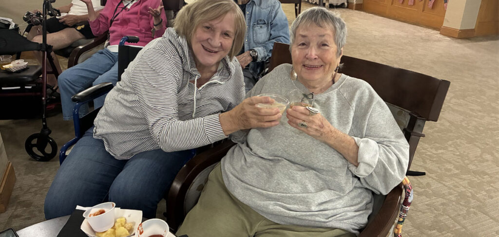Two people seated together at a community gathering, holding small cups and posing for a photo with snacks on a nearby table.