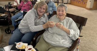 Two people seated together at a community gathering, holding small cups and posing for a photo with snacks on a nearby table.